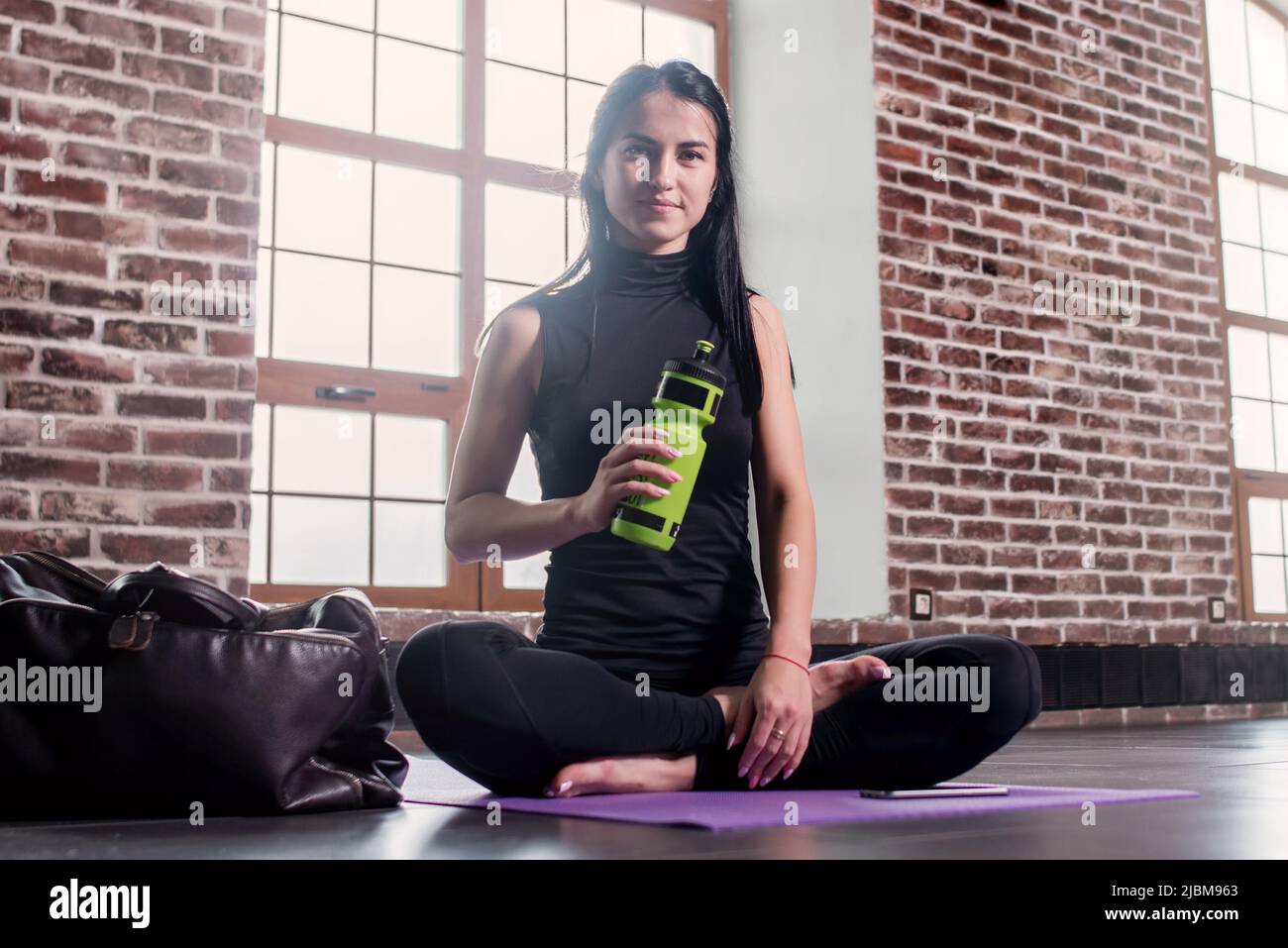 Portrait de la jeune femme caucasienne se reposant après l'entraînement tenant une bouteille de sport assise sur un tapis avec les jambes croisées dans la salle de gym avec intérieur loft. Banque D'Images
