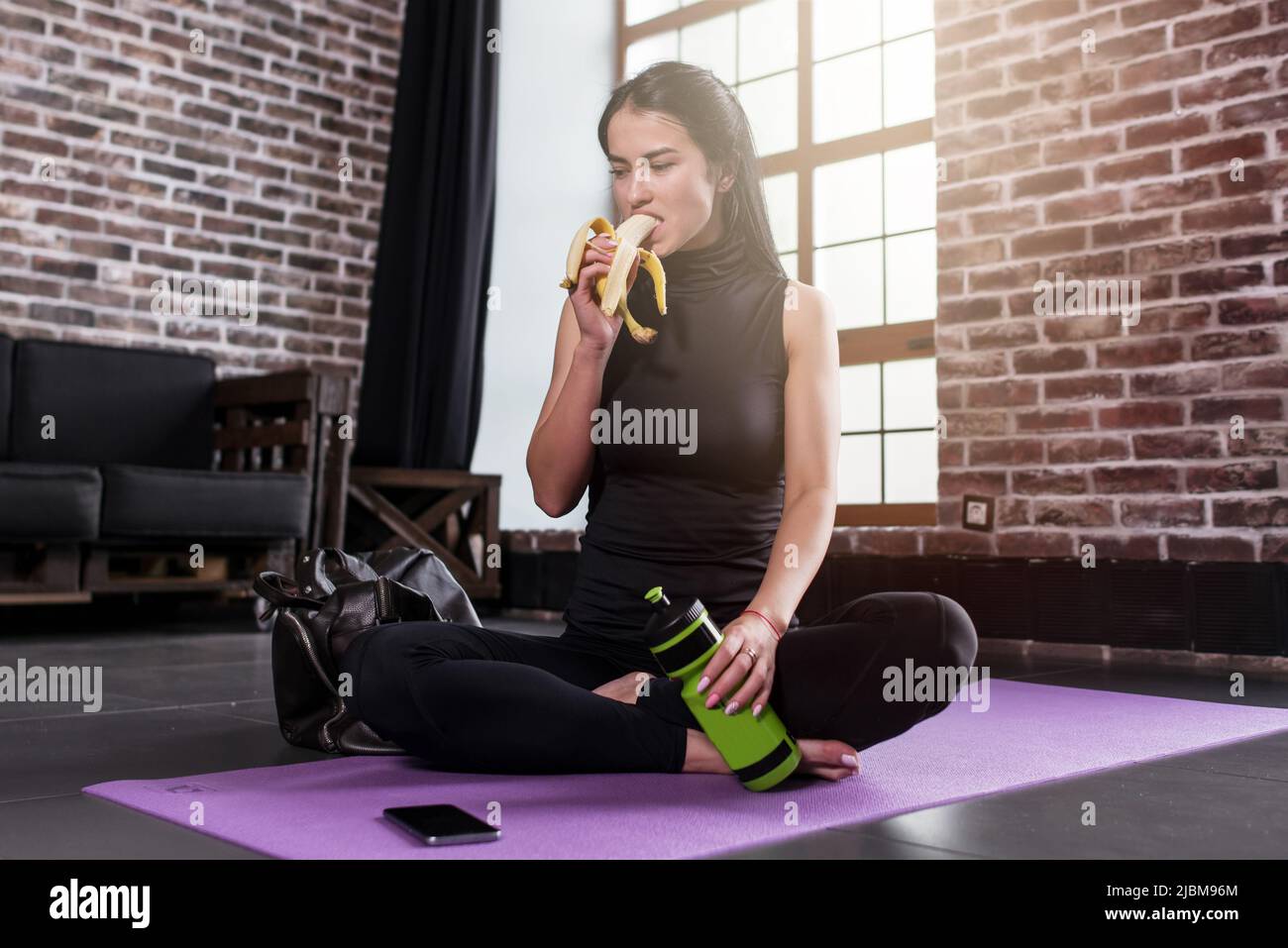 Portrait de la jeune femme caucasienne se reposant après l'entraînement tenant une bouteille de sport assise sur un tapis avec les jambes croisées dans la salle de gym avec intérieur loft. Banque D'Images