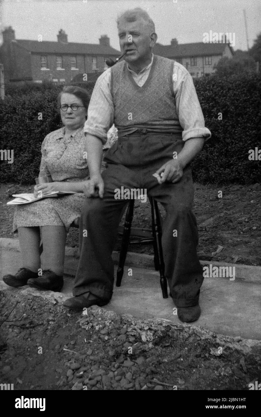 1940s, historique, sur une couche de concerete dans un jardin à l'arrière, un couple de travailleurs âgés assis dehors sur des tabourets, le gentleman assis sur un tabouret plus haut, d'où il semble assez grand que la dame, Angleterre, Royaume-Uni. Le gentleman fume un tuyau et porte un chandail sans manches sur une chemise sans col à manches enroulées. Banque D'Images