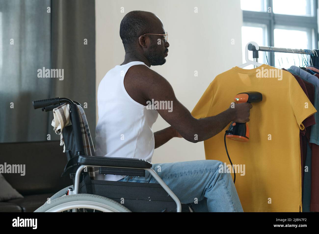 Jeune africain assis en fauteuil roulant à l'aide d'un défroisseur pour repasser les vêtements suspendus sur le rack Banque D'Images