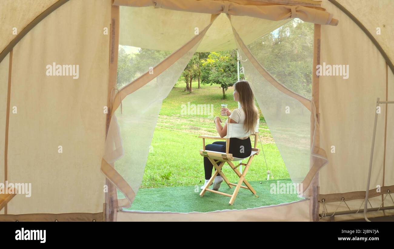 Concept d'éco-tourisme. Vue depuis la tente moderne sur une femme de voyage assise à l'extérieur de la tente moderne, boire de l'eau du verre, se détendre et profiter de la nature dans le parc sauvage Banque D'Images