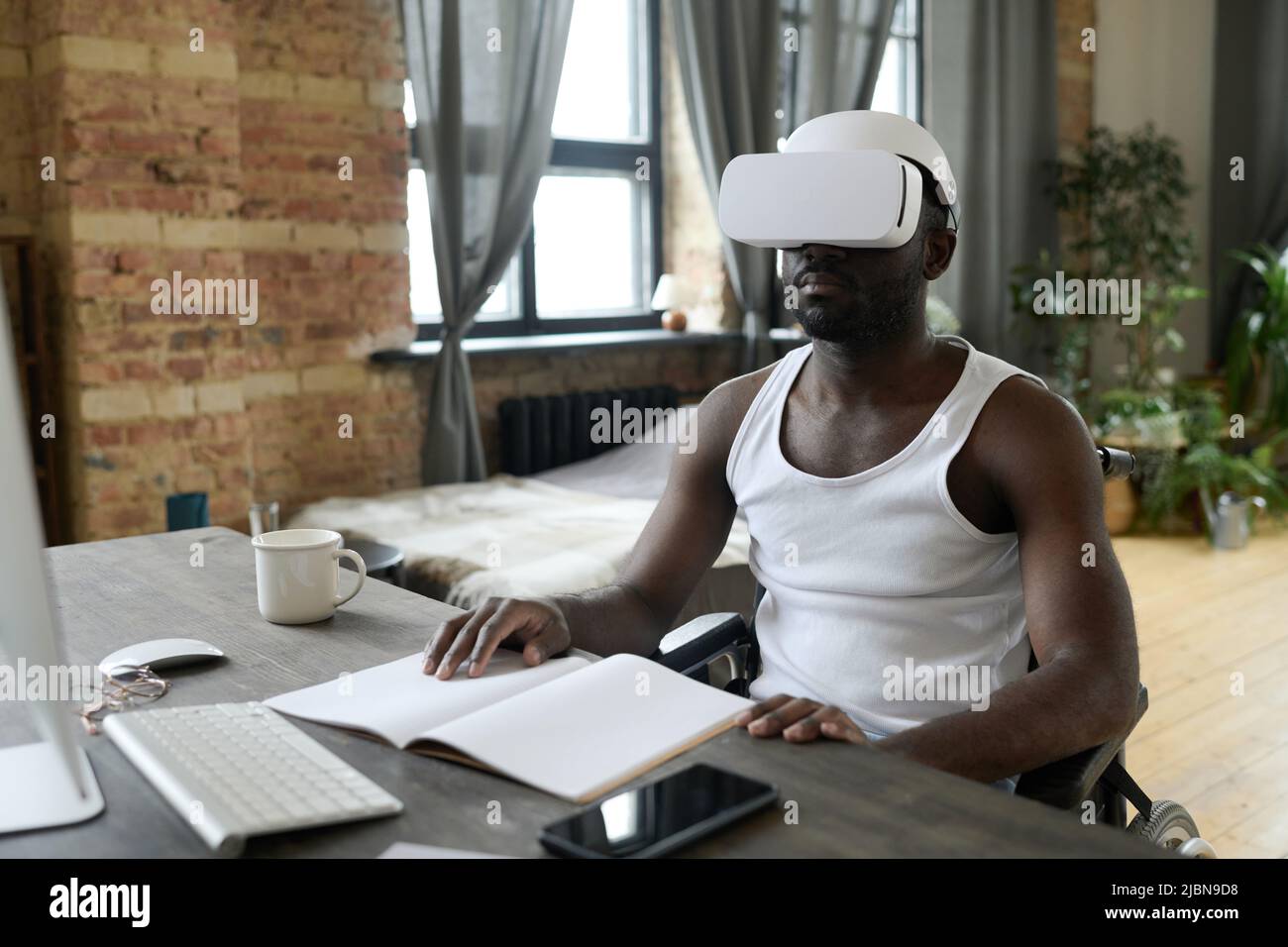 Jeune Africain handicapé étudiant en ligne à table à l'aide d'un casque de réalité virtuelle pendant la réadaptation à la maison Banque D'Images
