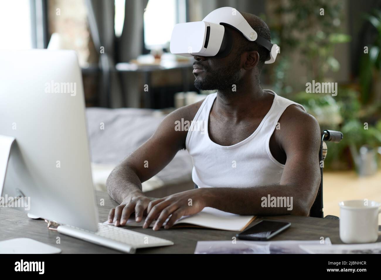 Jeune homme africain en réalité virtuelle lunettes assis à la table devant l'écran d'ordinateur et dactylographiant sur le clavier Banque D'Images