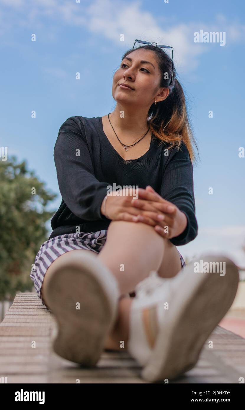 Une femme hispanique réfléchie, assise sur la place publique, regarde quelque chose en se reposant les deux mains sur son genou gauche. Banque D'Images