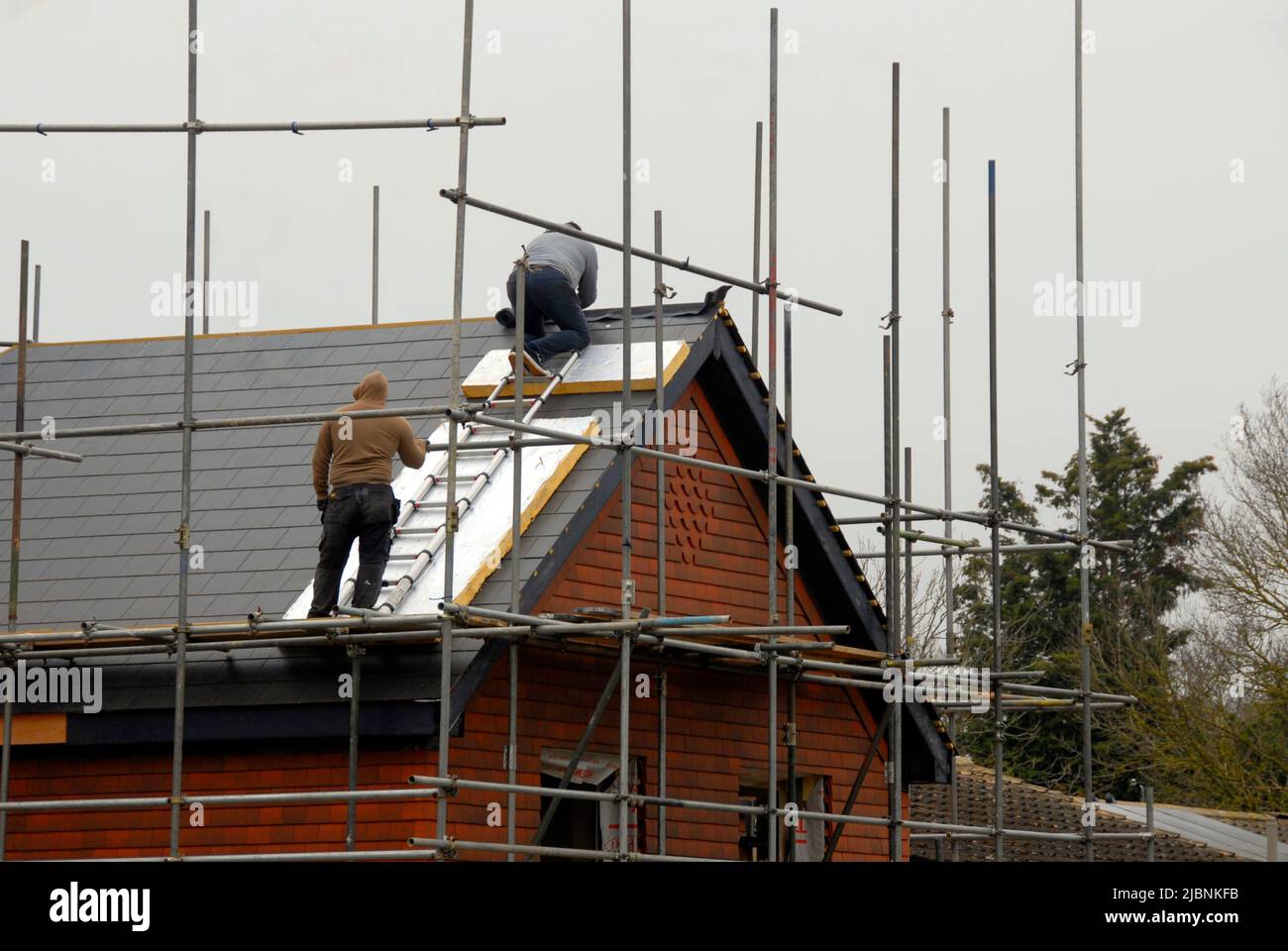 Deux hommes posant des carreaux de crête sur le toit d'une maison neuve à l'aide d'une échelle et de matériaux isolants mis au rebut pour protéger le toit Banque D'Images