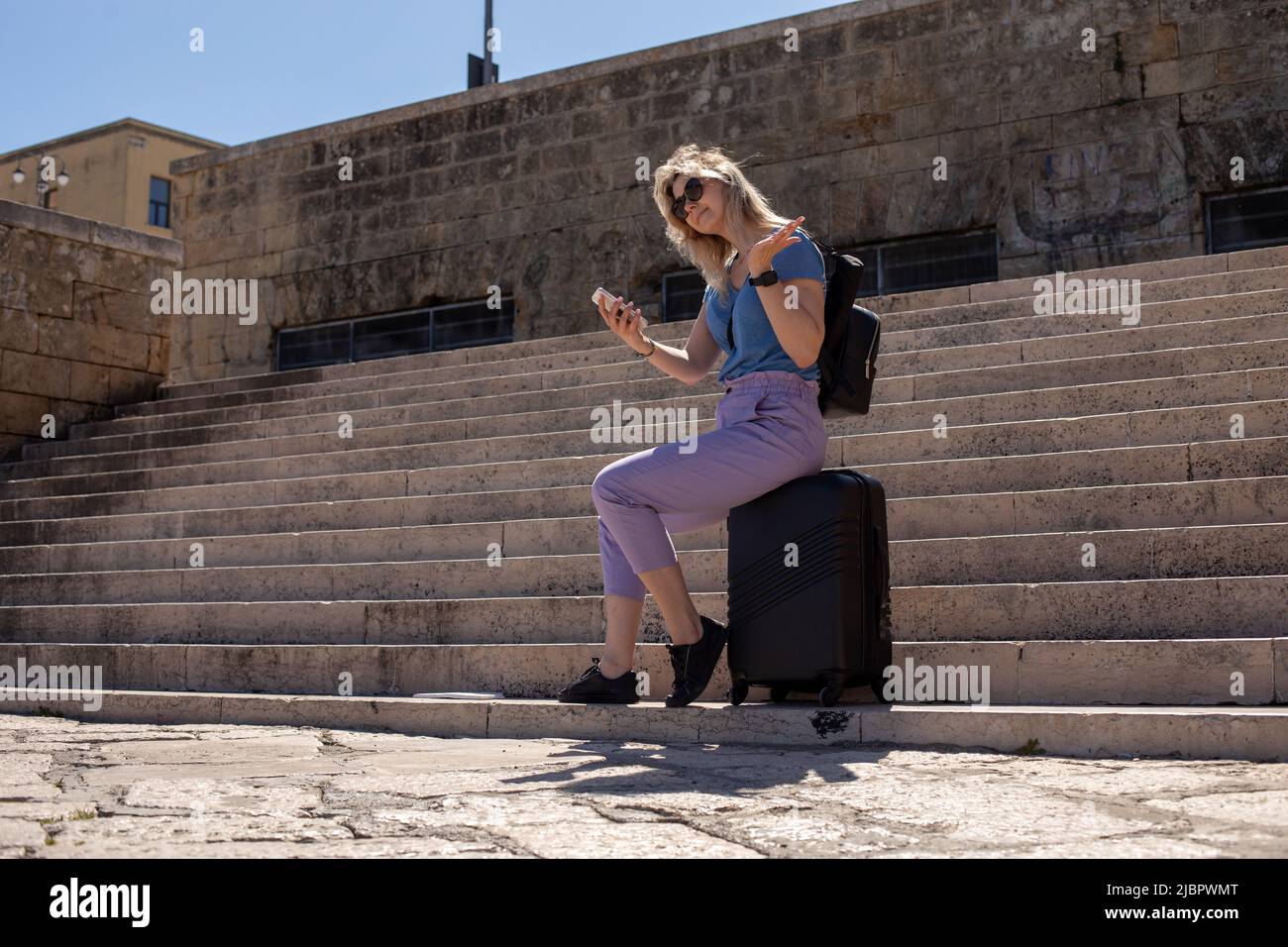 Jeune femme souriante assise sur une valise noire près des escaliers en béton de l'ancien bâtiment, tenant un smartphone, faisant des gestes. Banque D'Images