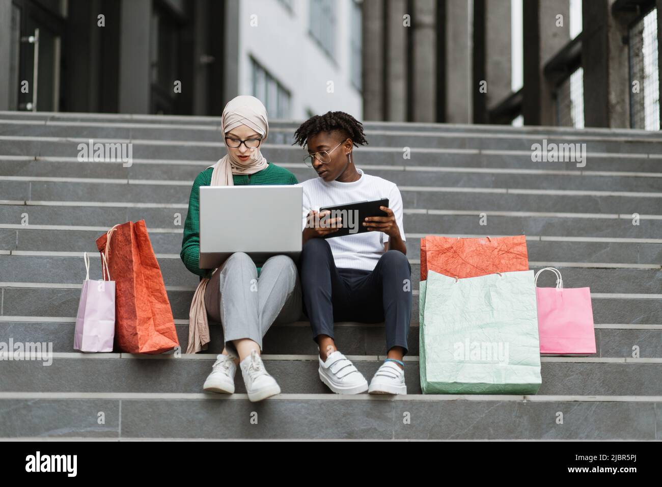 Portrait de mode plein air de deux jeunes femmes multiraciales dans des  vêtements élégants assis sur les marches d'un grand centre commercial et commander  des vêtements en ligne sacs de shopping colorés