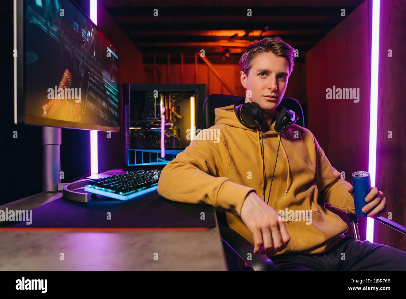Portrait d'un jeune joueur professionnel sérieux assis à un bureau avec un équipement de jeu, tenant une boisson énergétique, utilisant un PC puissant pour jouer à des jeux de tir vidéo en ligne. Chambre couleur néon, concept e-Sport Banque D'Images