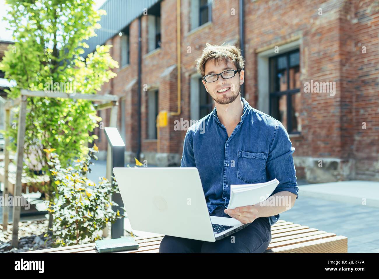 Portrait d'un jeune enseignant à l'extérieur du campus universitaire, assis sur un banc avec un ordinateur portable et souriant Banque D'Images