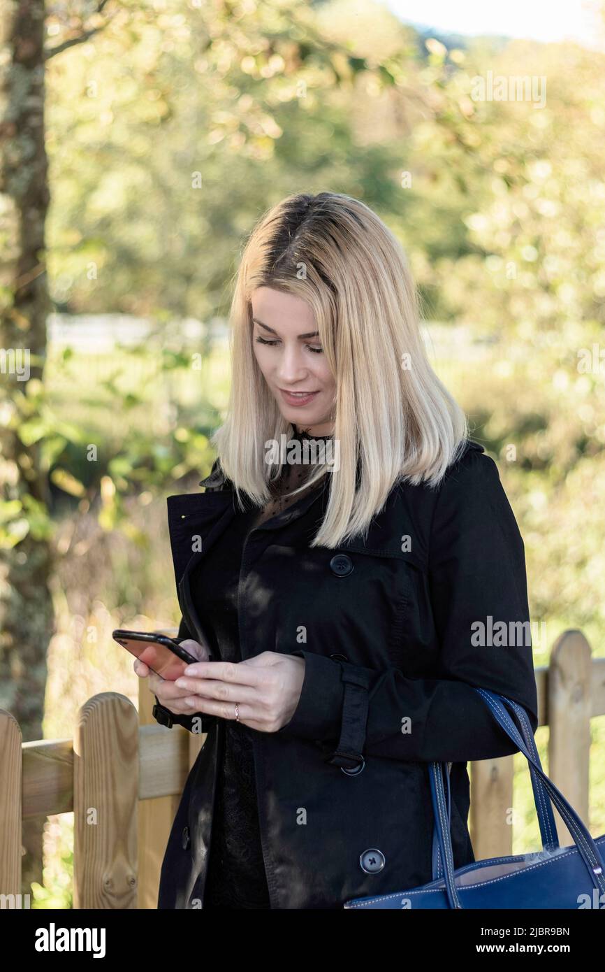 femme blonde utilisant le mobile dans un parc vêtu de noir et avec un sourire Banque D'Images