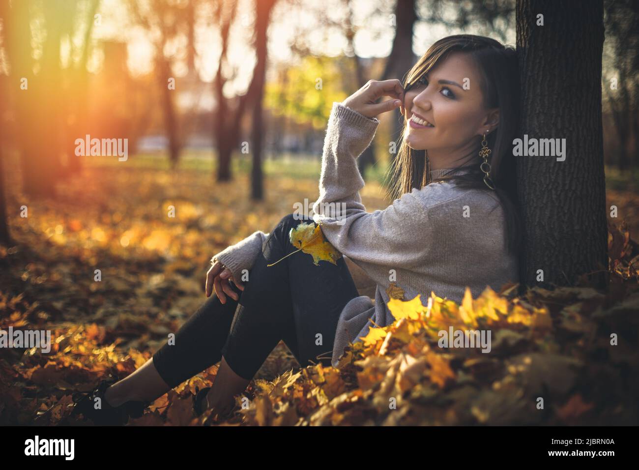 Jeune heureuse femme de course mixte assise près de l'arbre dans le parc d'automne au coucher du soleil regardant la caméra Banque D'Images