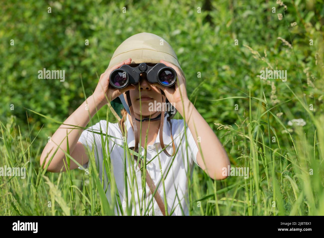 La préadolescente portant un casque de pième se cachant dans l'herbe regardant à travers des jumelles observant la nature estivale. Concept de découverte et d'aventure Banque D'Images