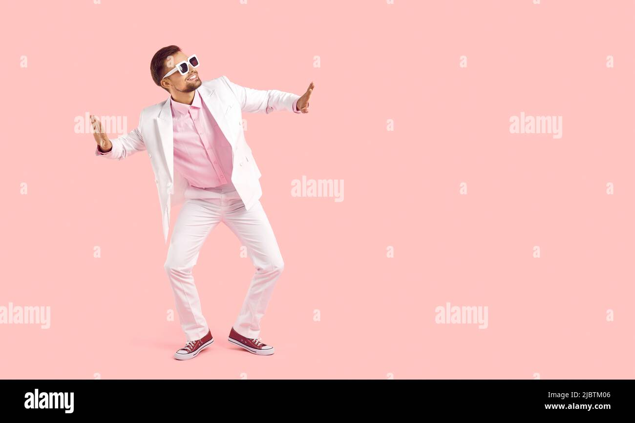 Joyeux jeune homme en costume blanc et lunettes de soleil dansant sur fond rose de l'espace de copie Banque D'Images