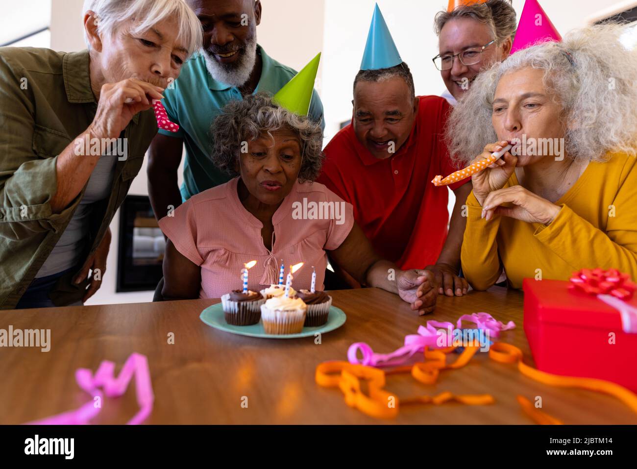 Des amis multiraciaux chantent et soufflent des cornes de fête tandis que la femme âgée soufflait des bougies d'anniversaire Banque D'Images