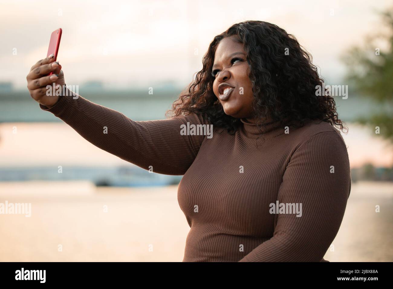 Curieusement confiant moderne multiculturel peau sombre femme en robe prenant le selfie au téléphone, posant, grimace. Corps positif Banque D'Images