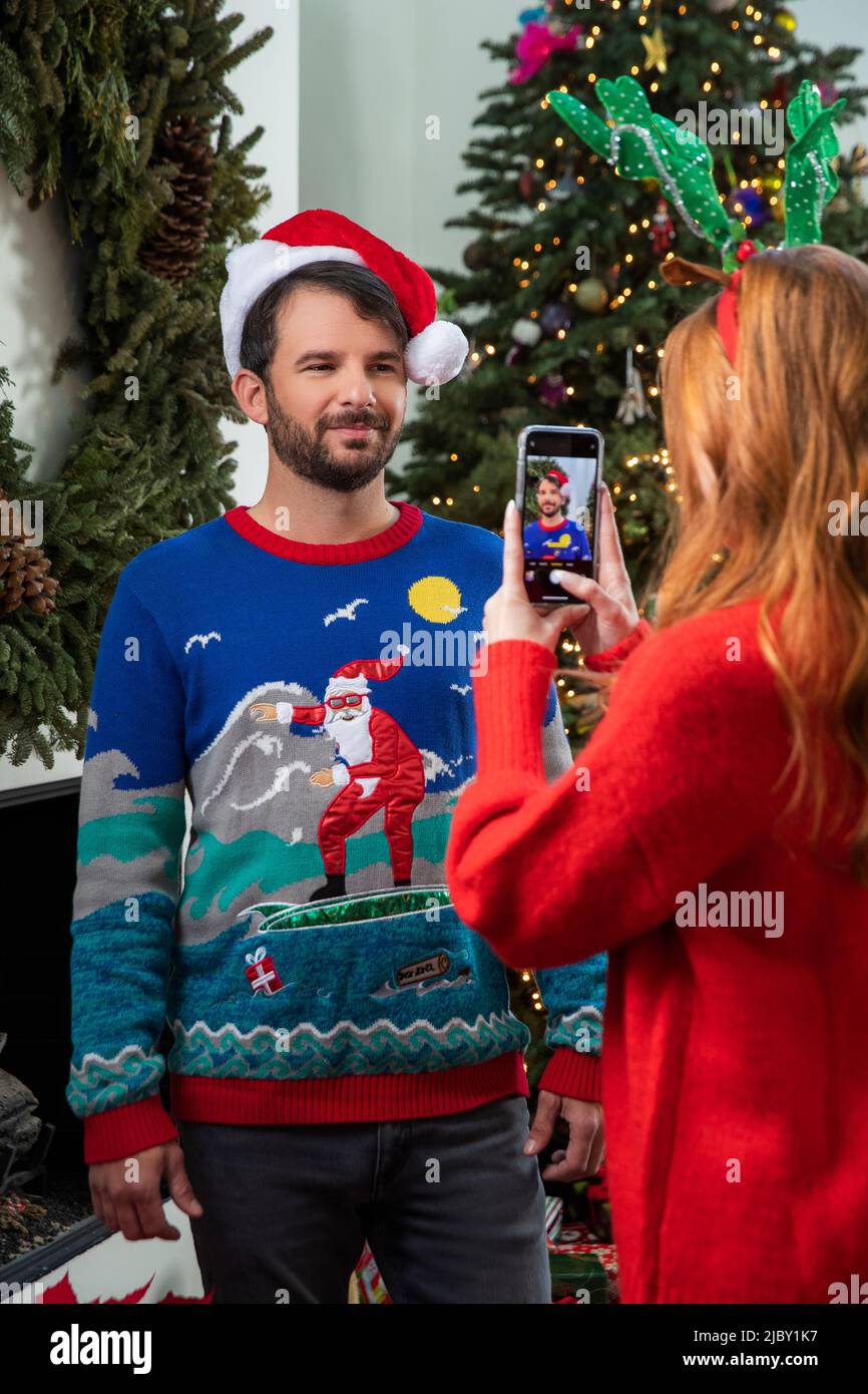 Heureux couple caucasien prenant des photos devant l'arbre de Noël et la couronne, portant des chandails de Noël. Banque D'Images