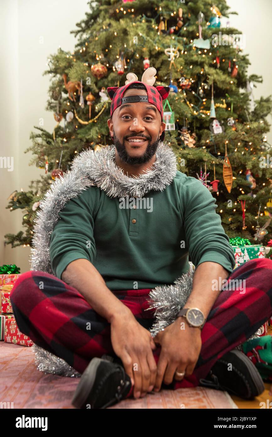 Portrait d'un Africain américain assis devant un arbre de Noël dans son pyjama décoré de guirlande argentée Banque D'Images