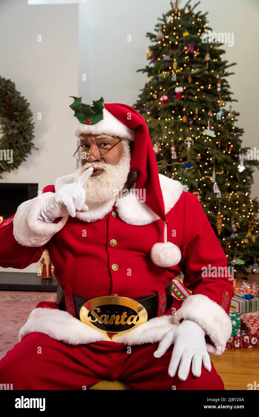 Le Père Noël afro-américain assis devant l'arbre de Noël avec son doigt jusqu'à la bouche se précipitant à la caméra. Banque D'Images