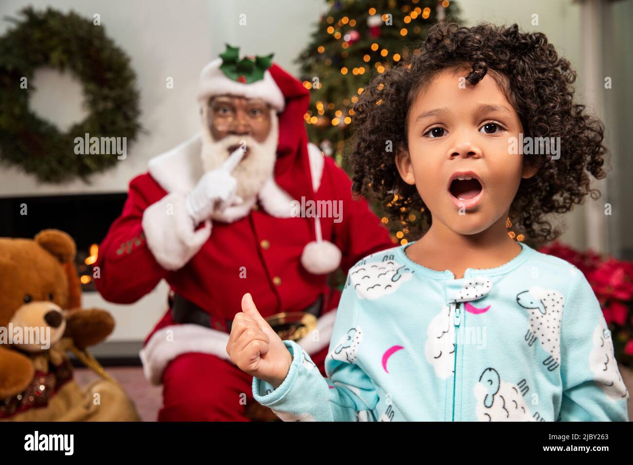 Portrait d'une petite fille debout devant l'arbre de Noël avec le Père Noël se faufilant dans l'arrière-plan. Père Noël noir. Banque D'Images