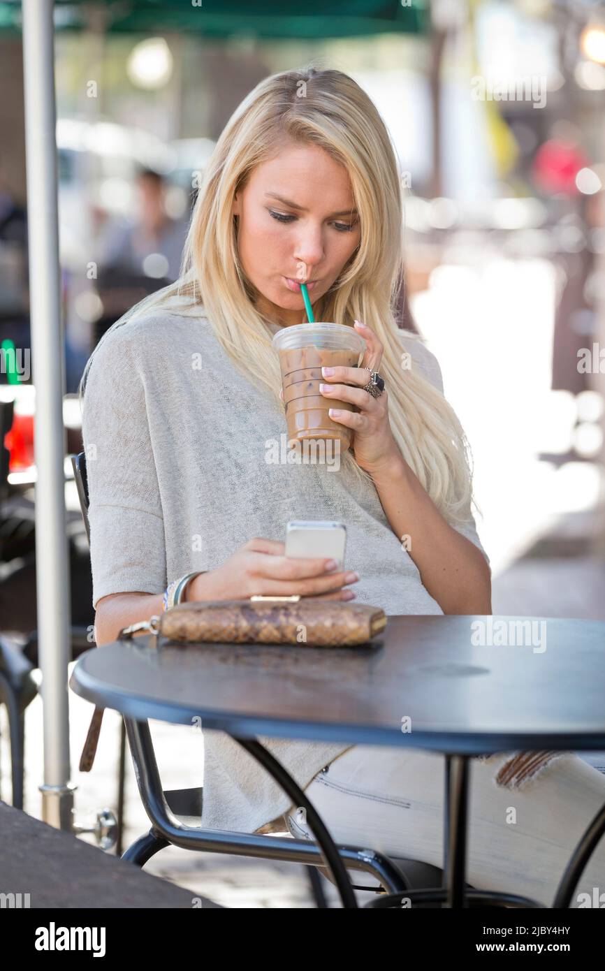Une jeune femme assise à l'extérieur dans un café au téléphone buvant du café glacé. Banque D'Images