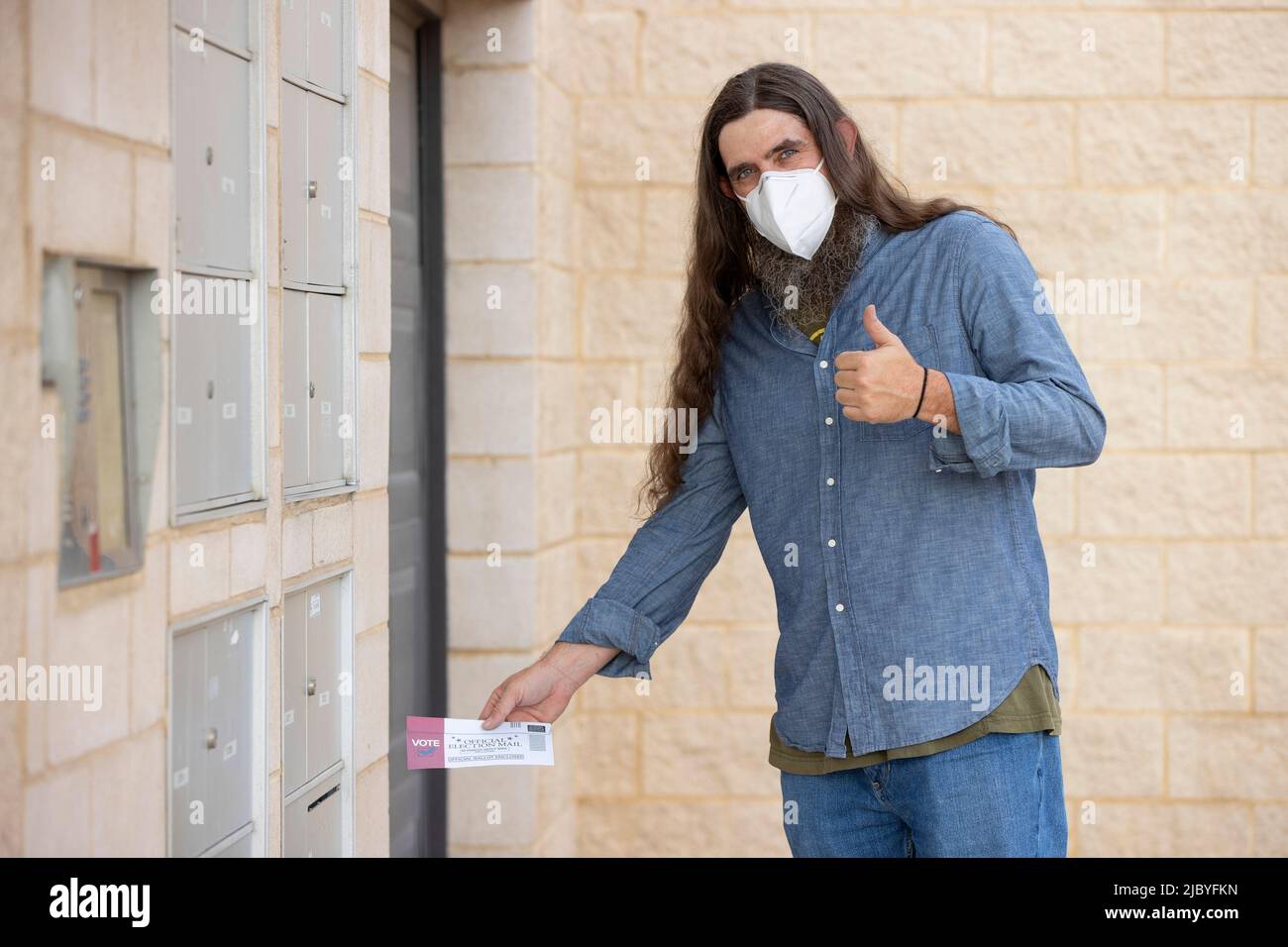 un homme aux cheveux longs et à la barbe complète portant un masque et mettant le courrier électoral officiel dans le bulletin de vote dans la fente de courrier sortant de son immeuble Banque D'Images