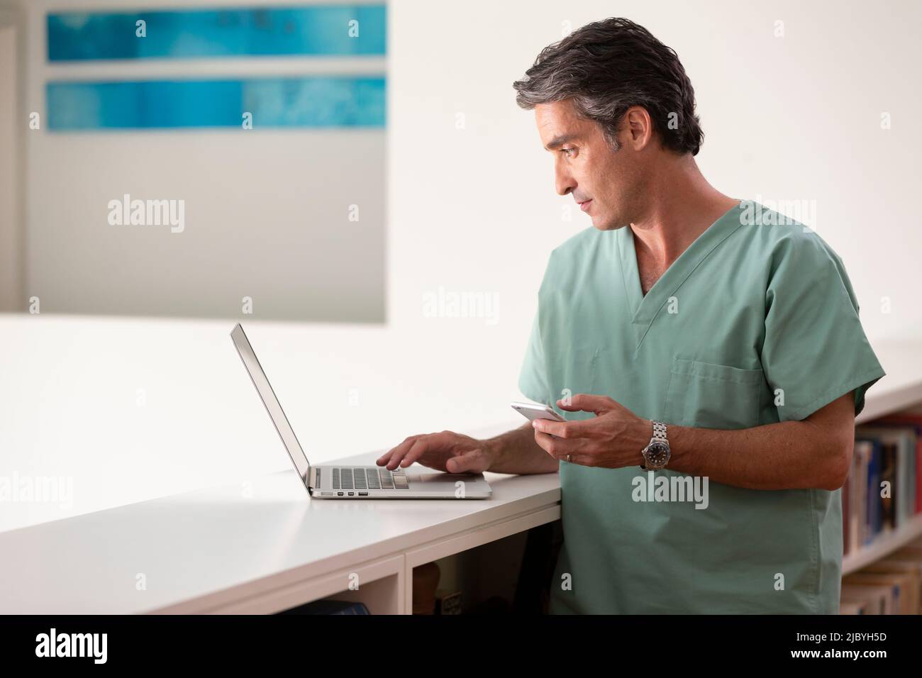 Hispanique Homme médecin dans sa maison pratiquant la téla-médecine, en utilisant l'ordinateur portable et le téléphone cellulaire Banque D'Images