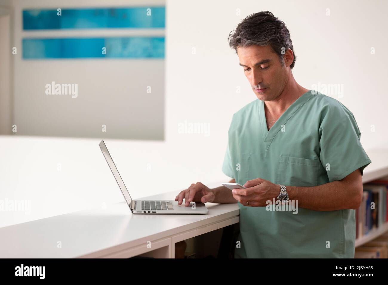 Hispanique Homme médecin dans sa maison pratiquant la téla-médecine, en utilisant l'ordinateur portable et le téléphone cellulaire Banque D'Images