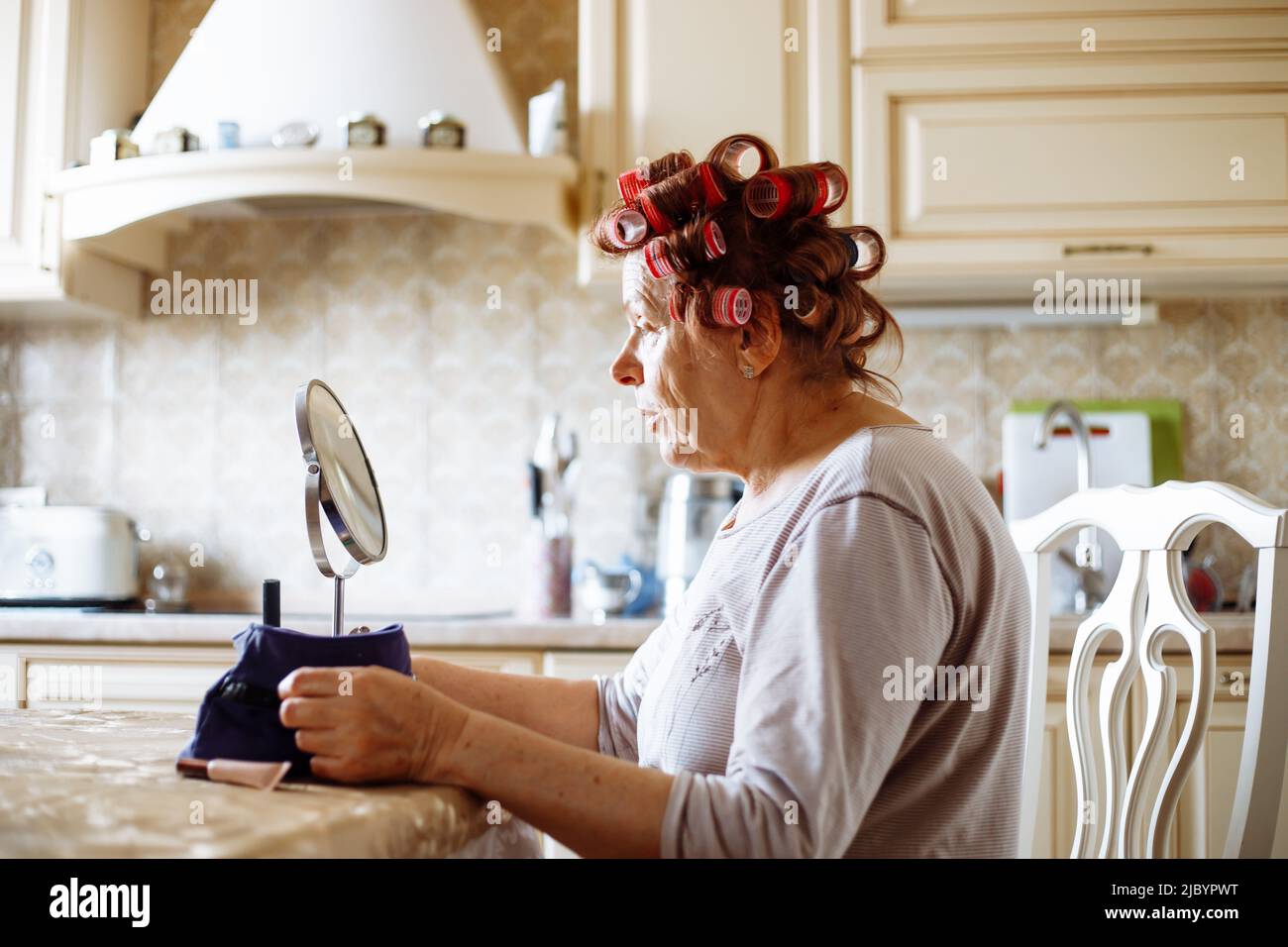 Vue latérale d'une femme âgée appliquant des bigoudis en plastique rose, assise à une table dans une cuisine beige, regardant le miroir. Banque D'Images