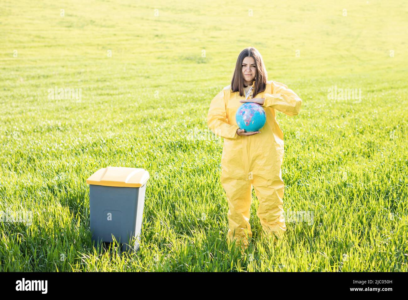 Une femme en costume de protection jaune se tient au milieu d'un champ vert et tient un globe entre ses mains, à côté d'une poubelle. Prenez soin de la planète, enregistrez le Banque D'Images