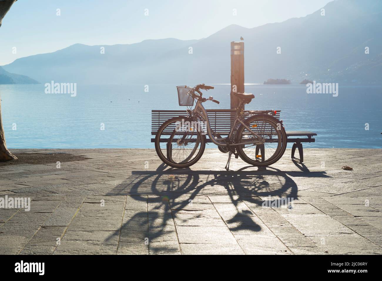 Bicycle penchée sur un banc sur le front de mer sur le lac alpin majeur avec la lumière du soleil et la montagne à Ascona, Suisse. Banque D'Images