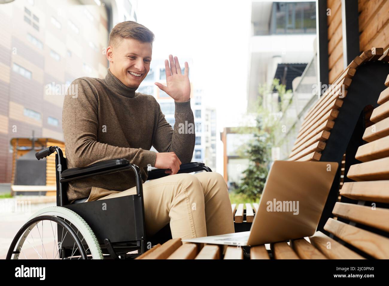 Joyeux jeune homme en chandail assis en fauteuil roulant à l'extérieur et en agitant la main tout en parlant via l'application de vidéoconférence sur ordinateur portable Banque D'Images