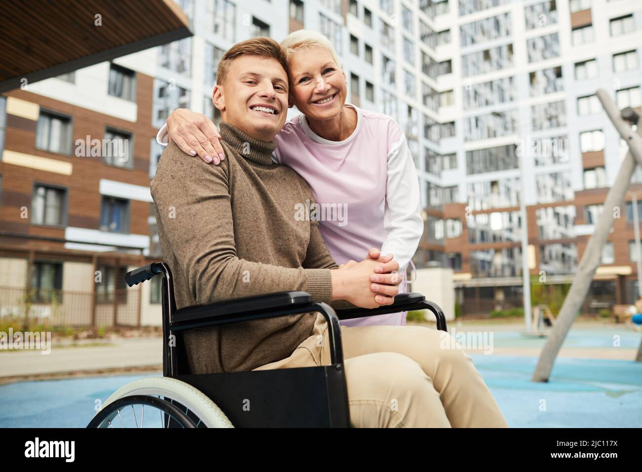 Portrait d'une infirmière positive et qualifiée qui embrasse un homme attaché à un fauteuil roulant alors qu'ils marchent sur une cour à proximité d'immeubles à appartements Banque D'Images