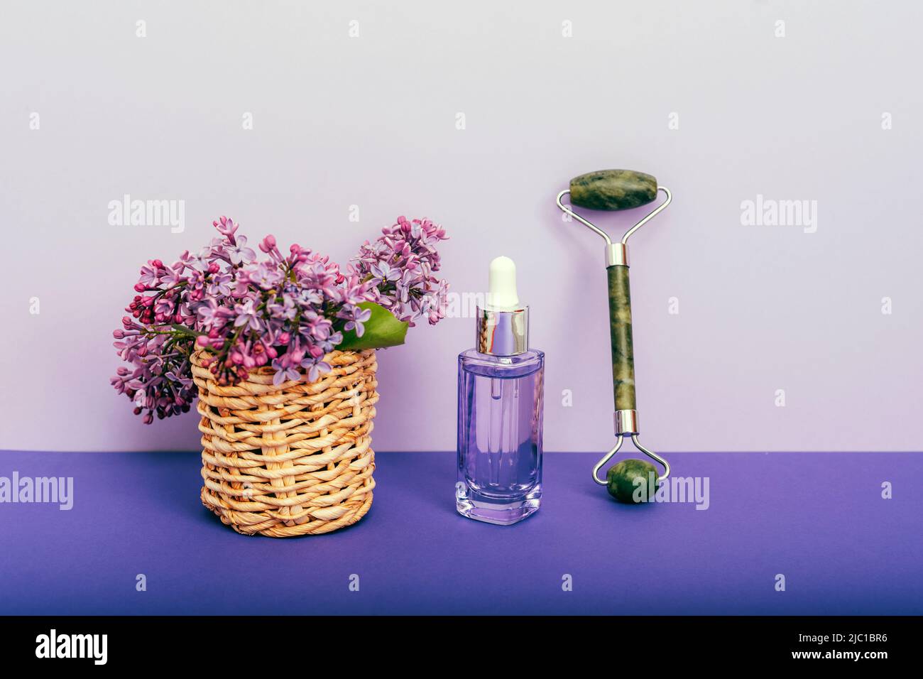 Bouteille de sérum cosmétique, rouleau facial jade et fleurs lilas sur table mauve. Cosmétiques naturels, concept de soins de la peau. Banque D'Images