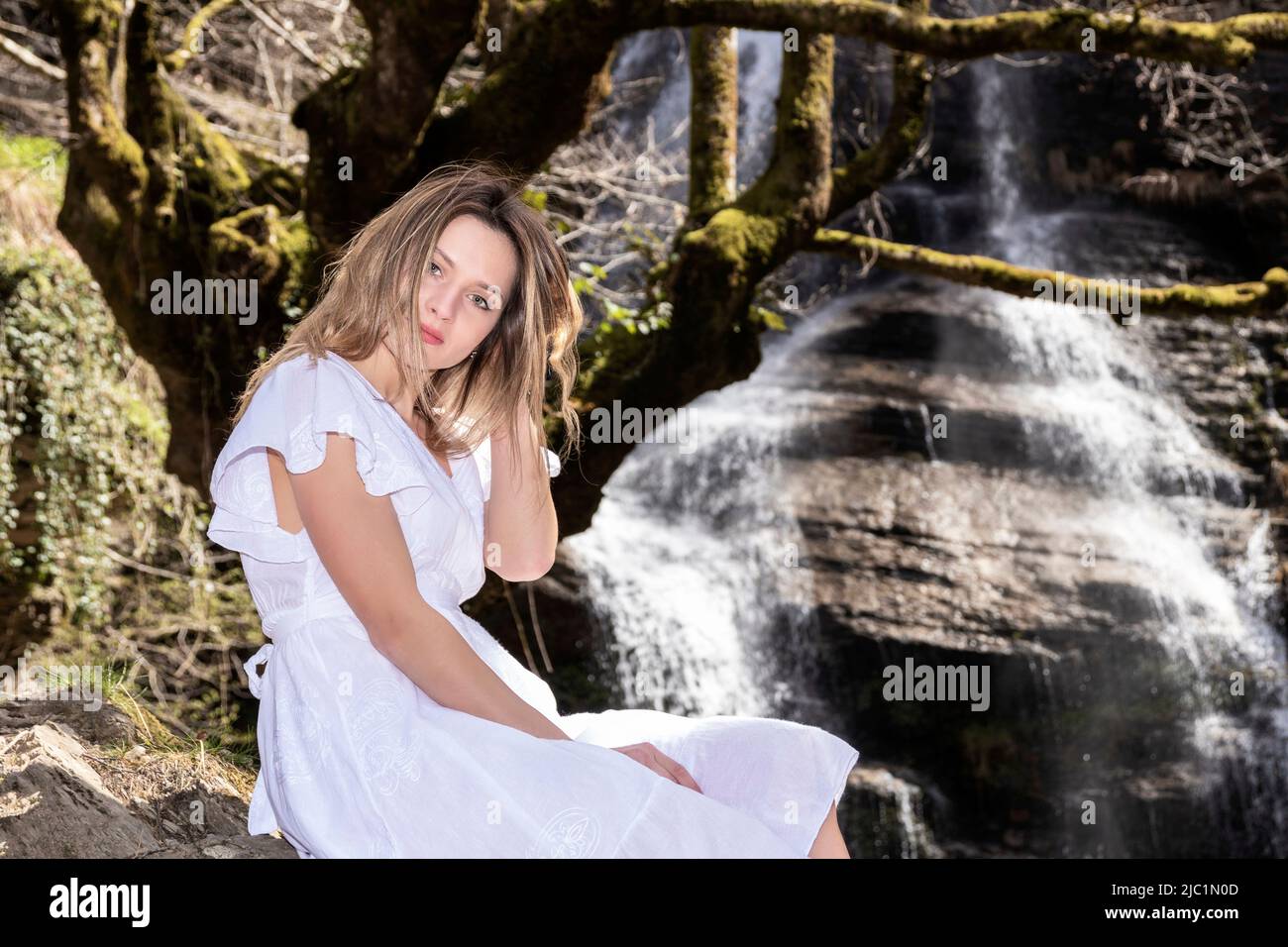 femme blonde en robe blanche assise dans la forêt devant une cascade Banque D'Images