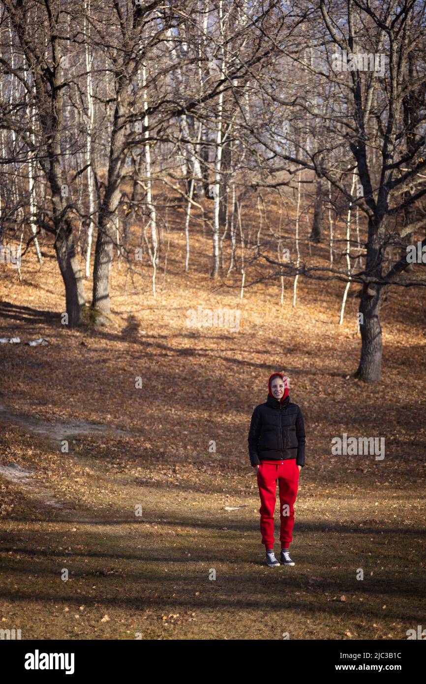 Une jeune femme blaieuse debout avec les mains dans les poches quelque part dans la forêt avec de grands arbres en arrière-plan et des feuilles sèches sur le sol regardant l'appareil photo allumé Banque D'Images