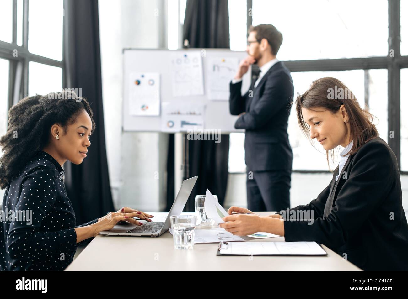 Des collègues féminins, une afro-américaine et une femme caucasienne, concentrés et prospères, sont assis à une table dans un bureau moderne, travaillent sur un projet, développent des start-up, communiquent avec les clients Banque D'Images