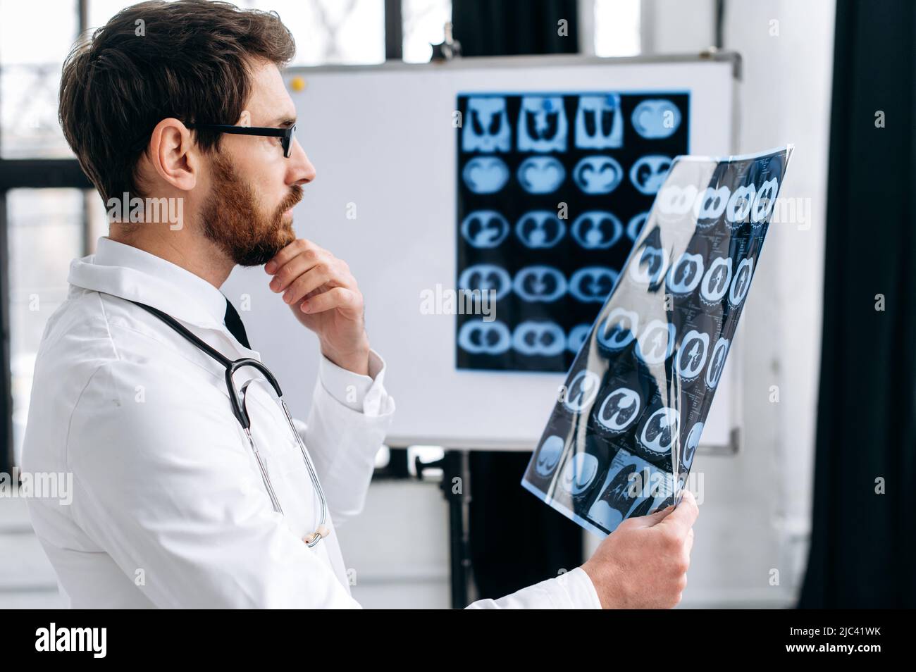 Focalisé bon médecin regardant la radiographie, le thérapeute mâle caucasien en uniforme médical blanc et des lunettes concentré de l'étude de la radiographie du patient, se tient dans le bureau médical. Concept de soins de santé Banque D'Images