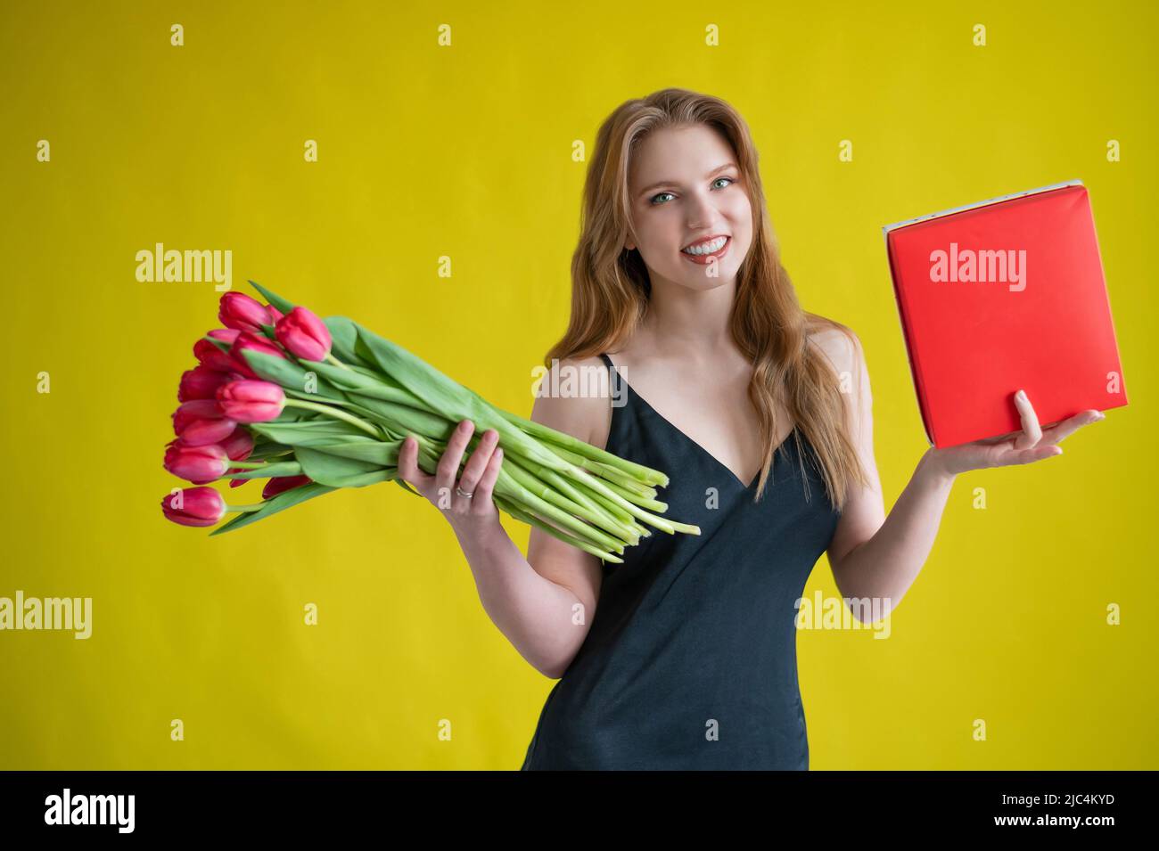 Femme avec un bouquet de tulipes rouges sur fond jaune.Une fille heureuse dans une robe noire tient une armful de fleurs et une boîte de chocolats.Cadeau pour Banque D'Images