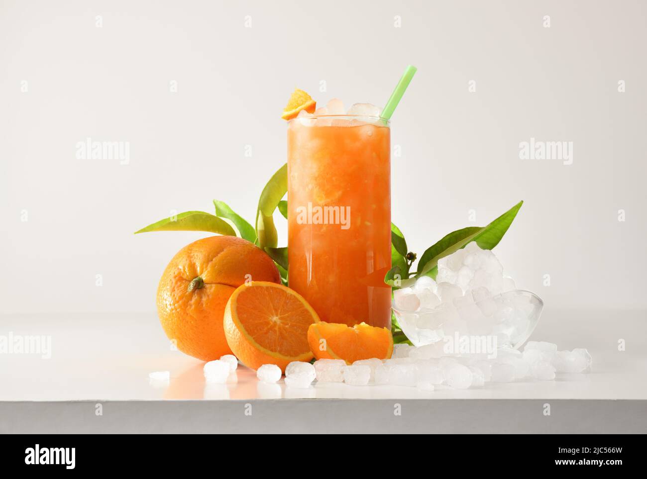 La slushie orange dans un grand verre avec des fruits et de la glace pilée autour de lui sur une table blanche avec un fond isolé. Vue avant. Composition horizontale. Banque D'Images