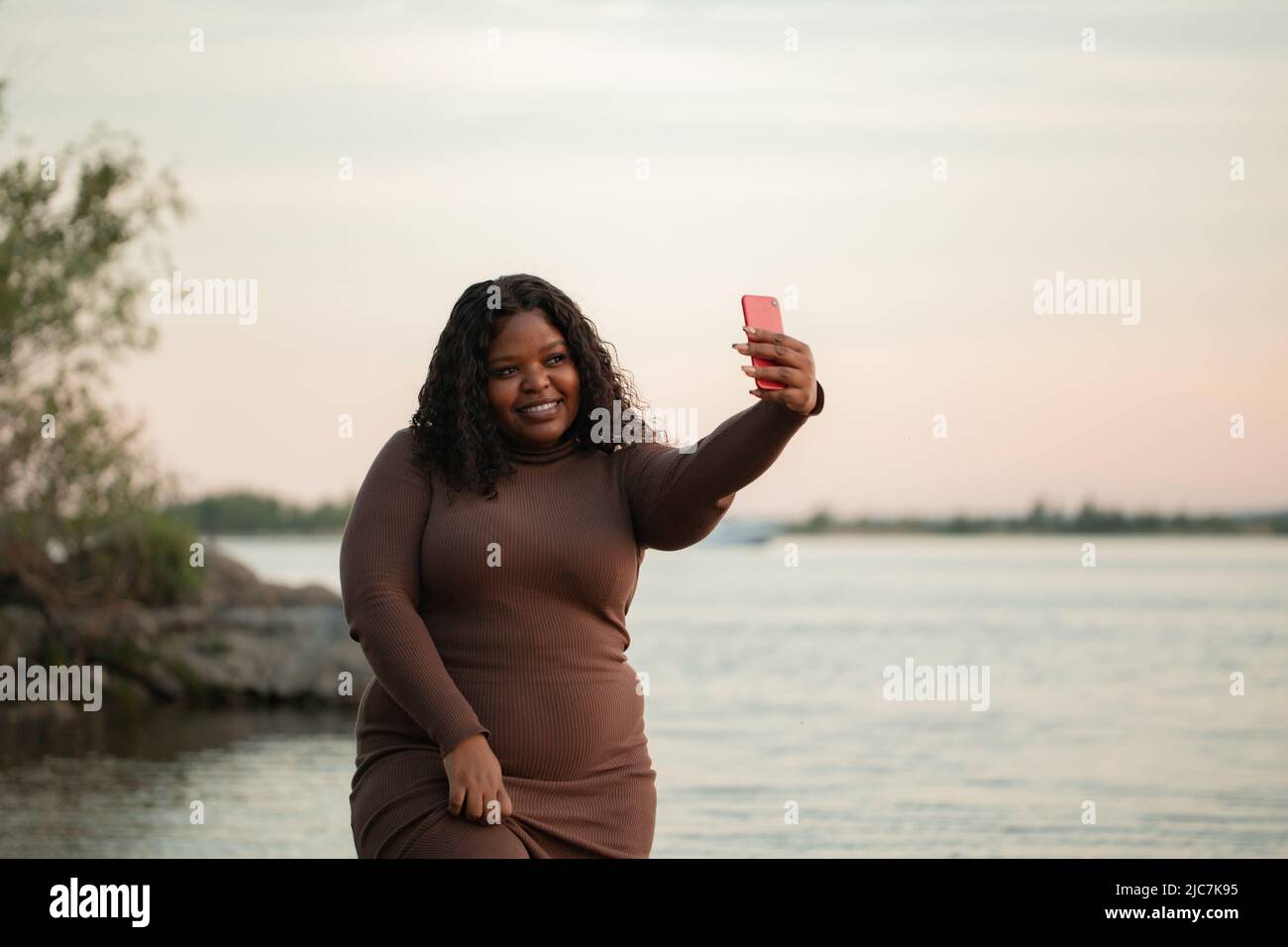 Jolie femme interraciale curieusement souriante avec afro en robe pendant les vacances d'été, promenade sur la plage, la mer ou la rivière prendre selfie Banque D'Images