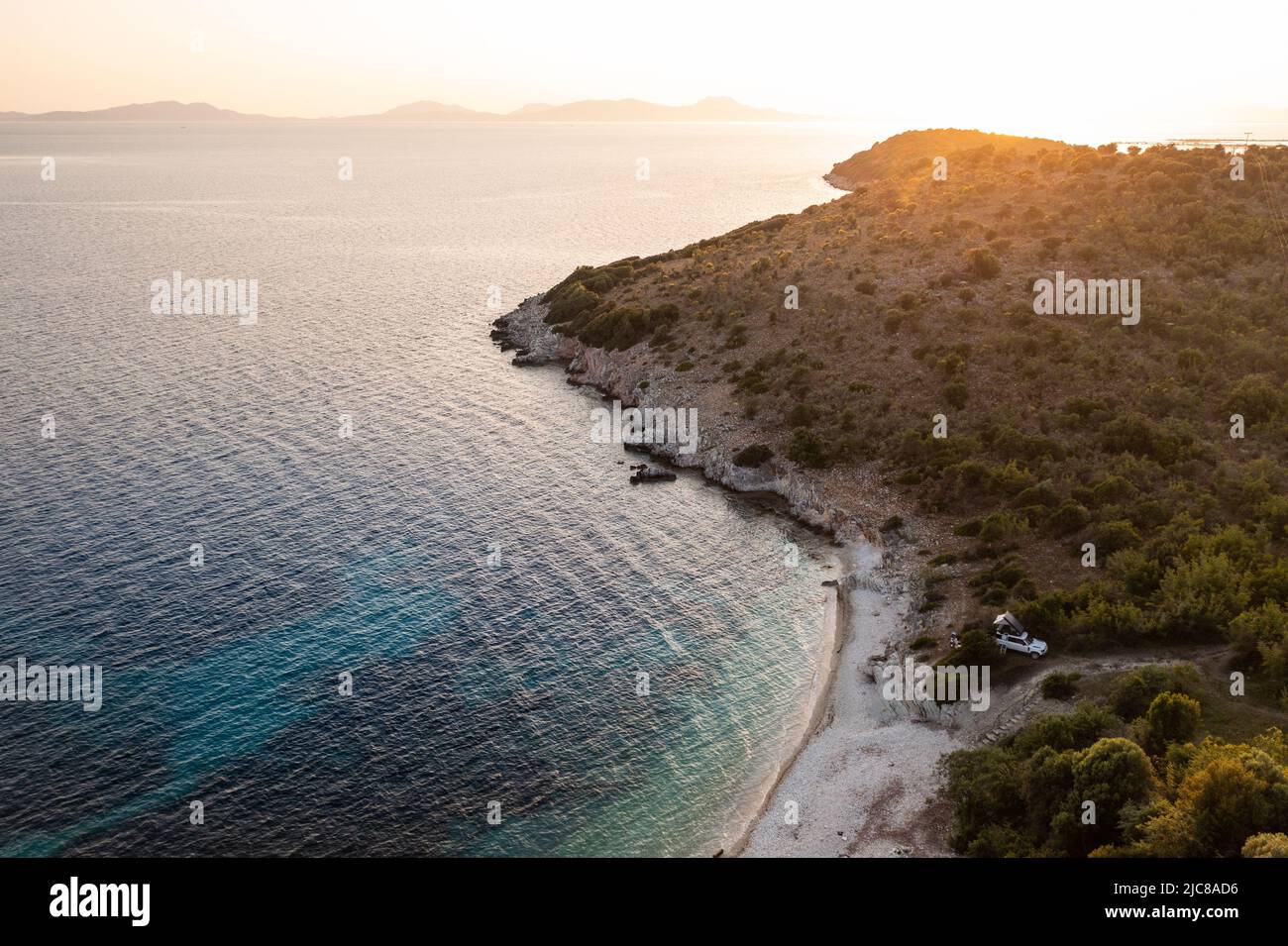 Voiture Offroad blanche avec tente sur le toit sur la côte grecque avec eau turquoise au premier plan et coucher de soleil en arrière-plan. Grèce. Banque D'Images