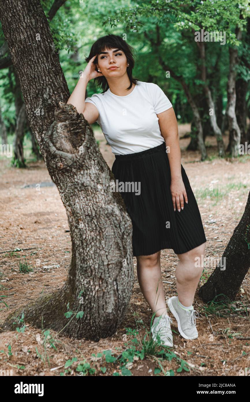 Une jeune fille aux cheveux noirs courts, un T-shirt blanc et une jupe noire dans les bois entre les arbres verts et l'herbe verte Banque D'Images