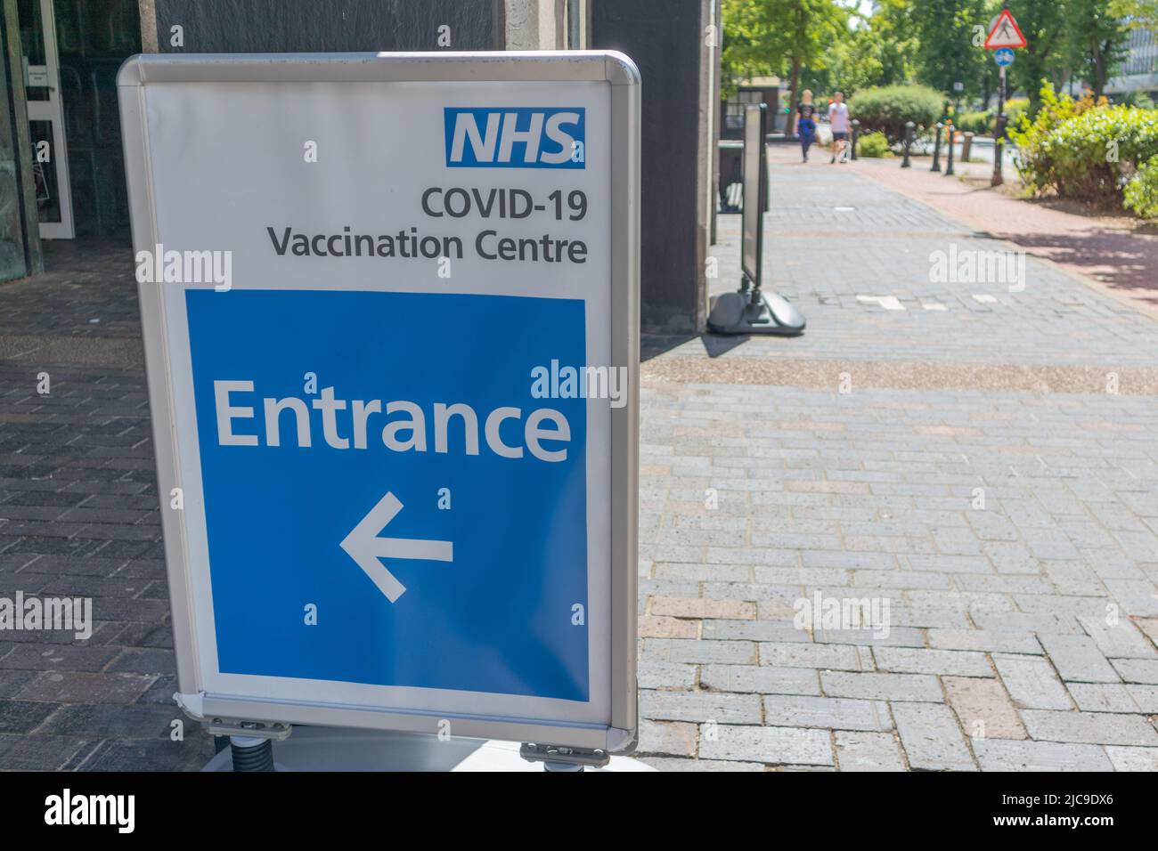 Southend on Sea, Royaume-Uni. 11 juin 2022. Un panneau directionnel pour un centre de vaccination COVID-19 du NHS placé sur un trottoir pavé de briques dans une zone urbaine britannique. Le panneau indique l'entrée avec une flèche pointant vers la gauche. Des piétons et un panneau de passage à niveau sont visibles à l’arrière-plan, soulignant l’infrastructure de santé publique pendant la pandémie. Une promenade dans le centre de vaccination Covid 19 à Southend. Penelope Barritt/Alamy Live News Banque D'Images