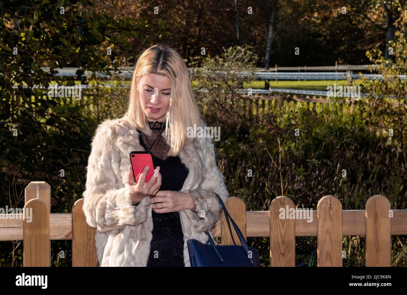 femme blonde regardant un mobile rouge dans un parc, vêtue d'un manteau blanc Banque D'Images