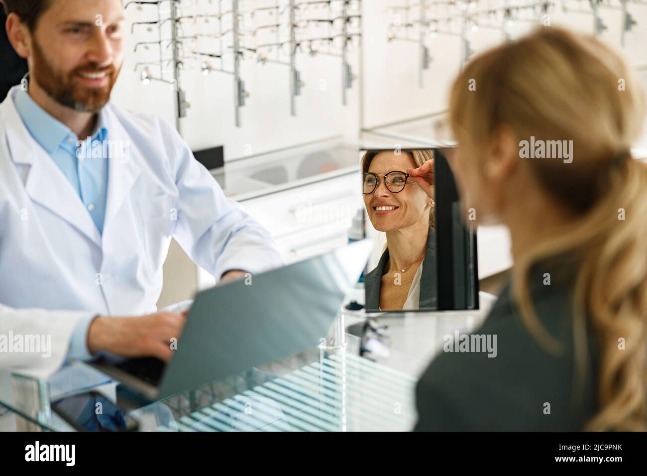 Optométriste masculin aidant la femme à choisir des lunettes dans le magasin optique montrant à son miroir Banque D'Images
