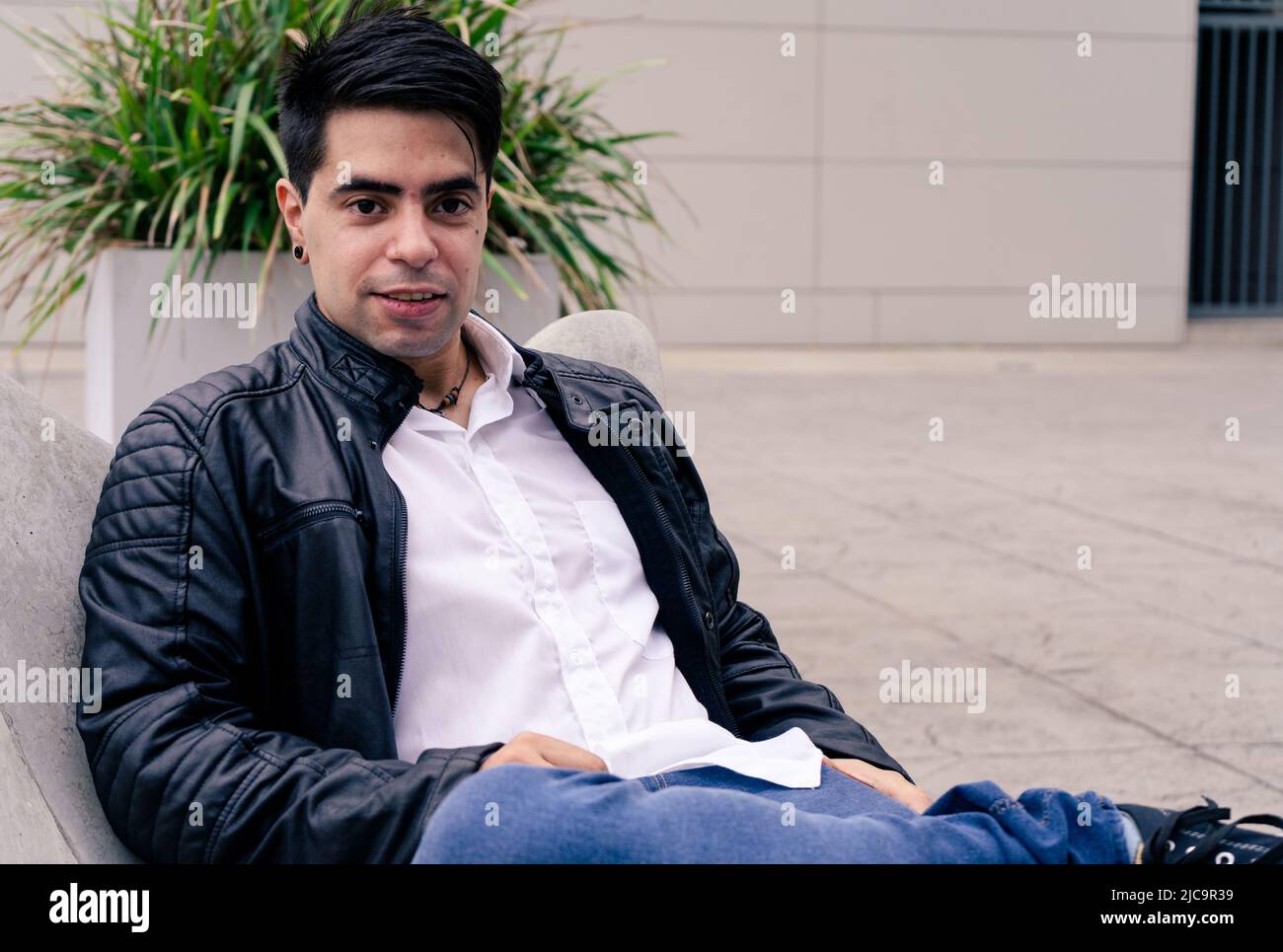 Bel homme Latino dans une chemise et une veste blanches, assis sur une chaise en ciment dans une place. Banque D'Images