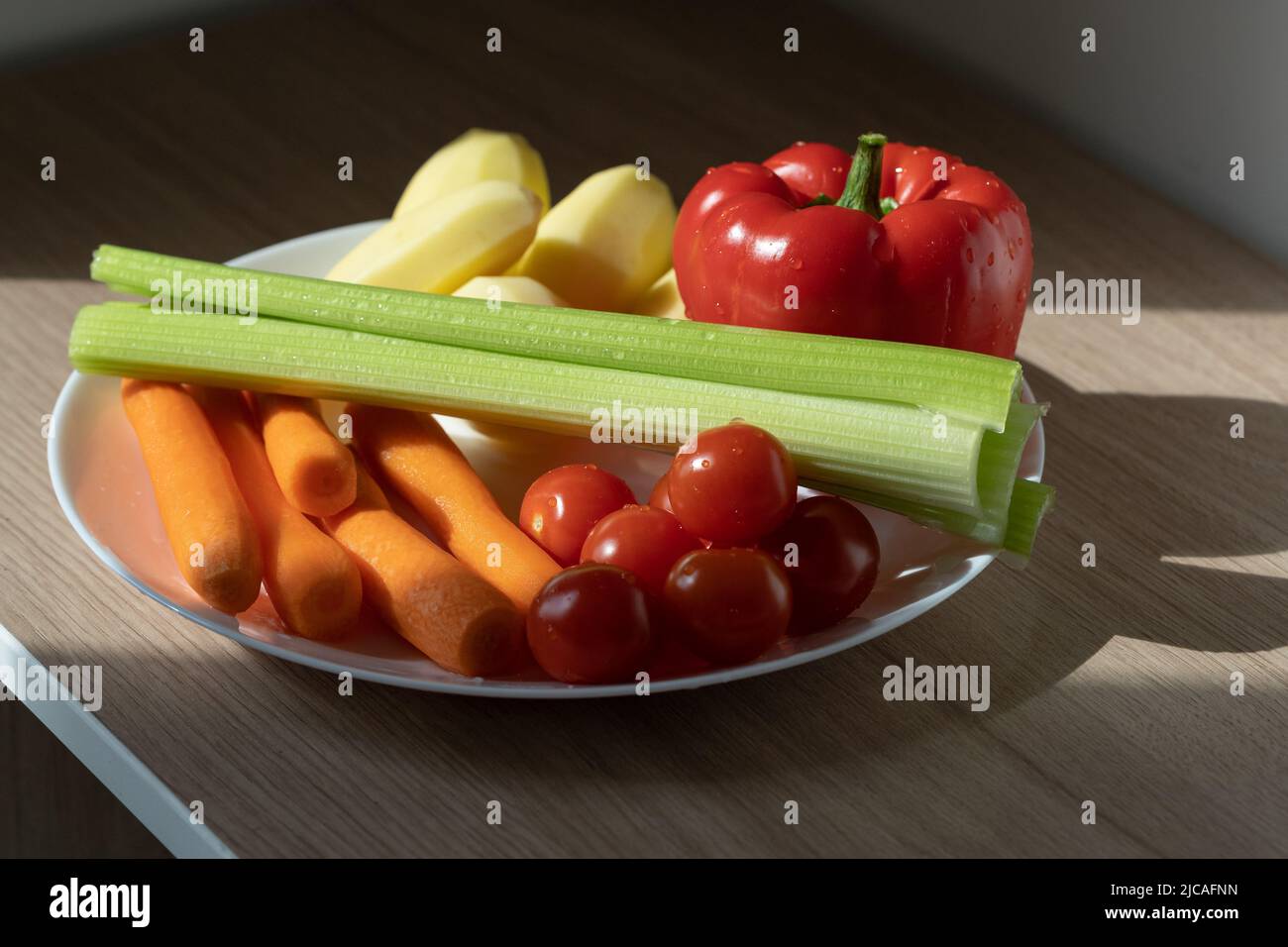 Assiette blanche avec tomates cerises, carottes, céleri, poivrons et pommes de terre au soleil sur table en bois. Banque D'Images