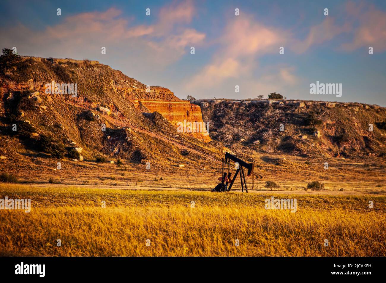 Tire-lait de puits d'huile dans la terre rouge de l'ouest de l'Oklahoma à l'heure d'or avec le ciel tôt du coucher du soleil et des bluffs en arrière-plan. Banque D'Images