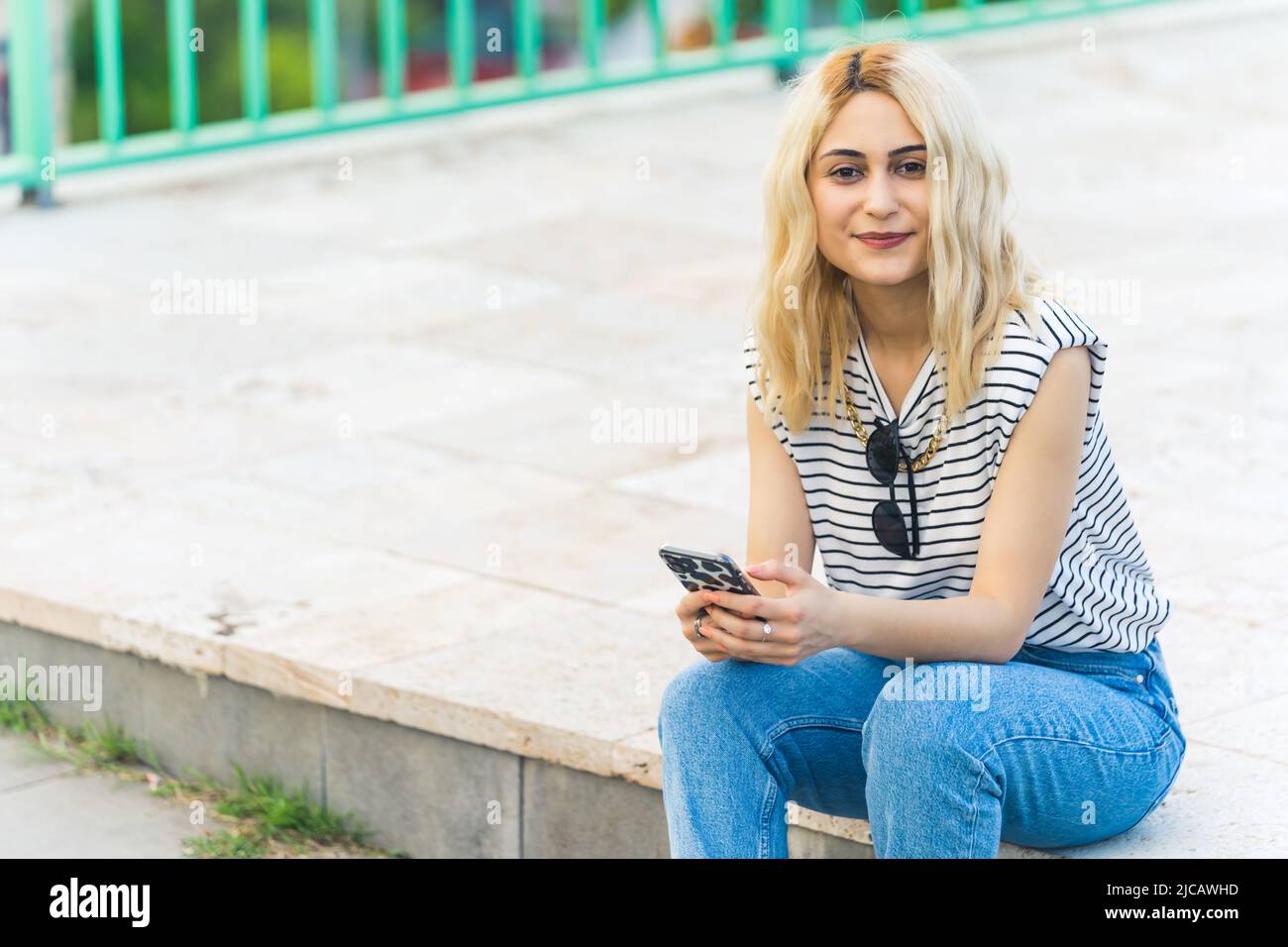 Prise de vue pleine et moyenne d'une jeune fille blanche blonde tenant son téléphone et souriant à l'extérieur. Photo de haute qualité Banque D'Images