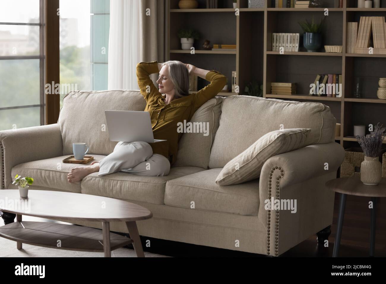 Une femme âgée sereine se reposant sur un canapé avec théière et ordinateur portable Banque D'Images